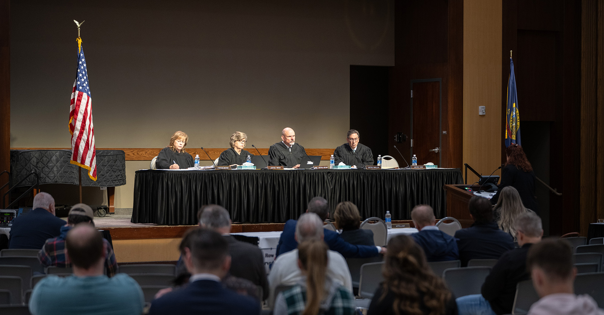 The Idaho Supreme Court Justices sit at a table on stage in an auditorium and listen to a lawyer as she delivers her oral arguments in a case. Fifteen people sit in rows of chairs and observe the court proceedings.